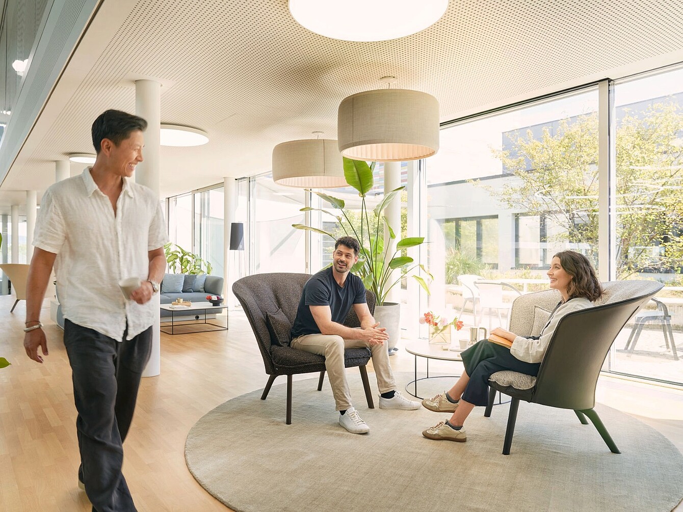 Two people sit in armchairs on a round rug in a bright room with large windows while a third person walks by.