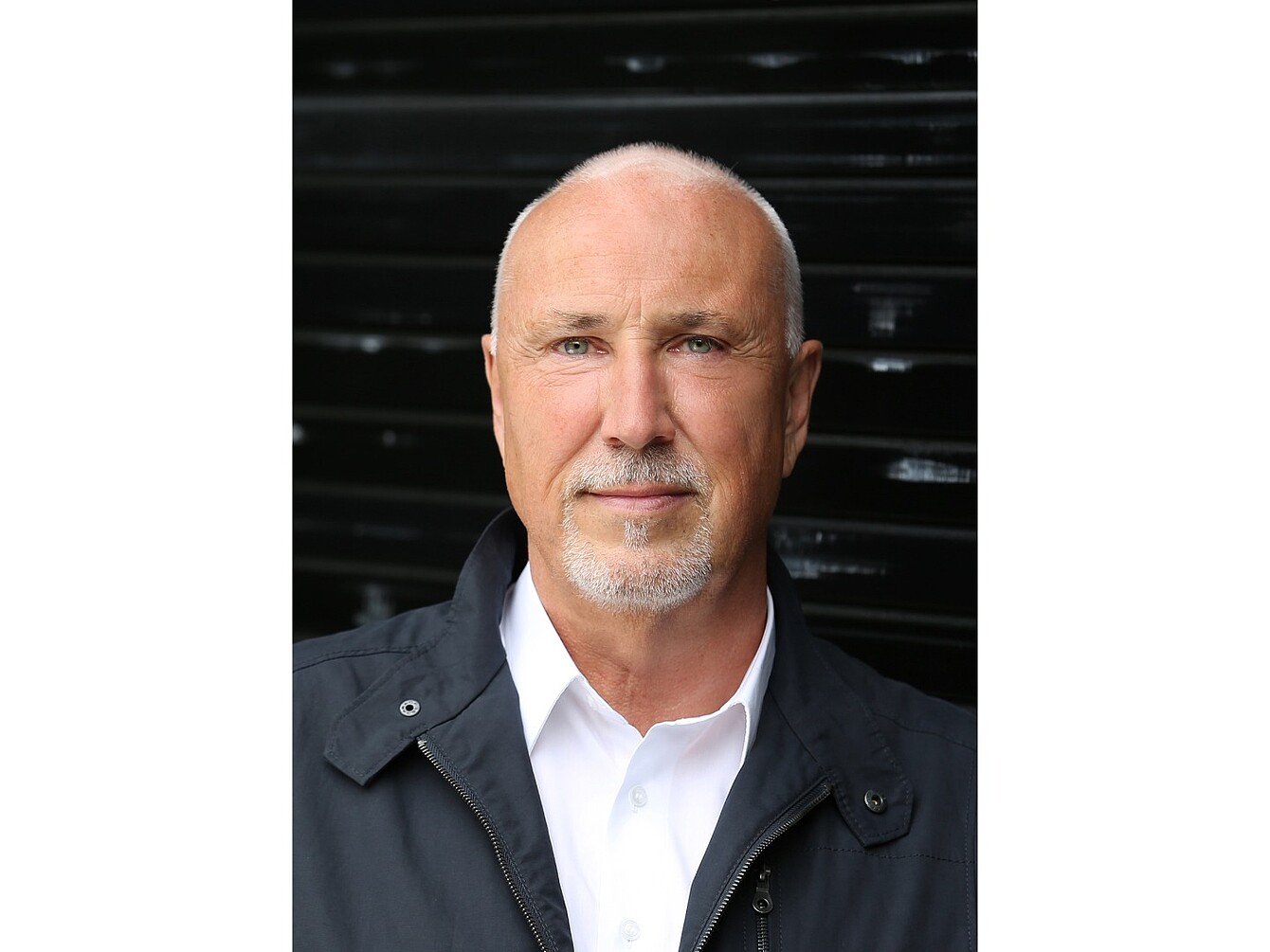 Man with short white hair (Jörg Bakschas) wearing a white collared shirt and dark jacket against a dark background.