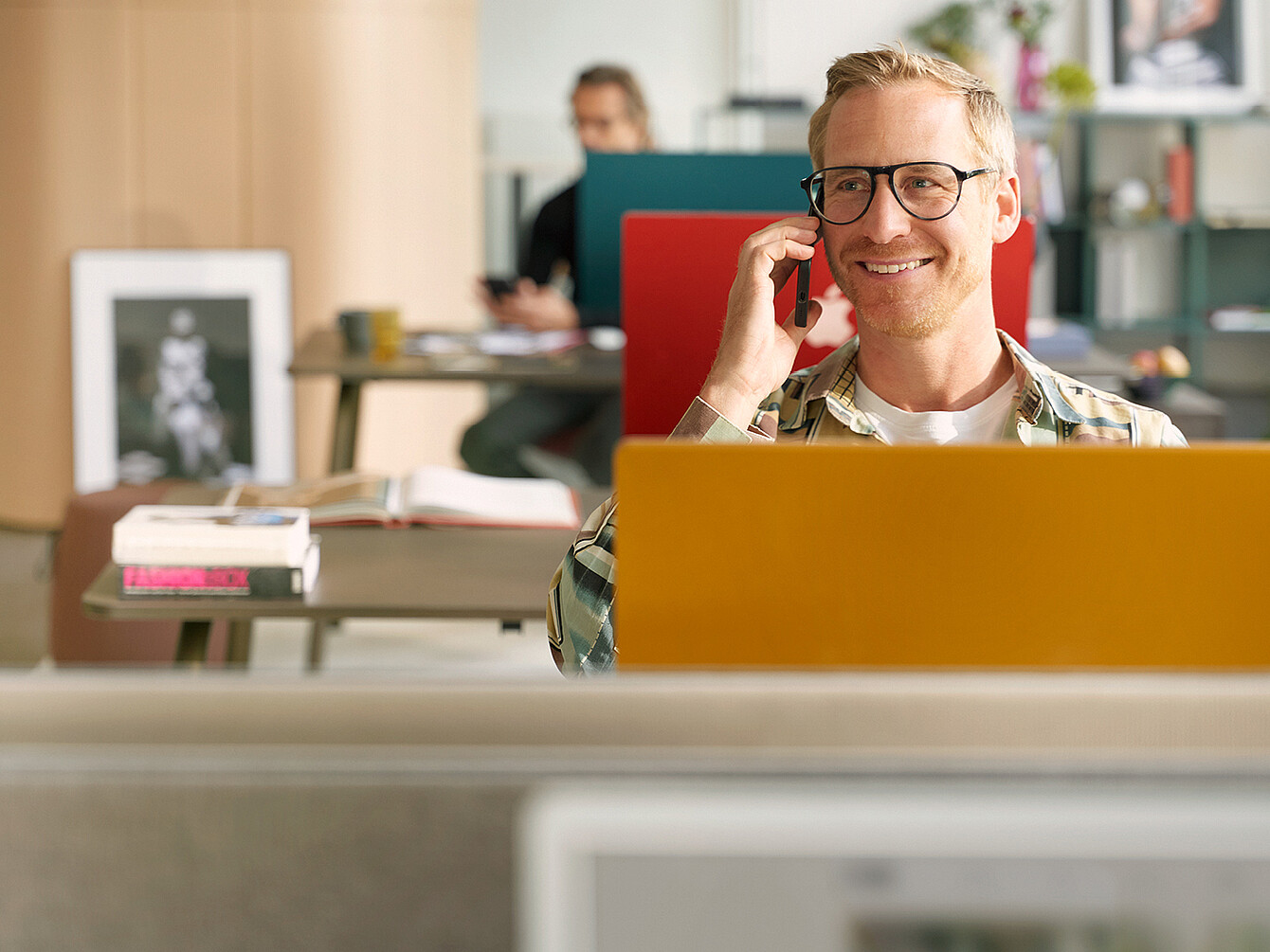 Uomo con gli occhiali seduto dietro un laptop giallo in un ufficio mentre parla al telefono con una persona che lavora a una scrivania sullo sfondo