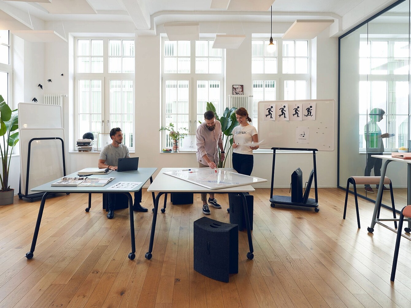 Three people collaborate in a bright office with large windows, standing and sitting around tables with laptops and papers, a whiteboard with simple human figure drawings is visible