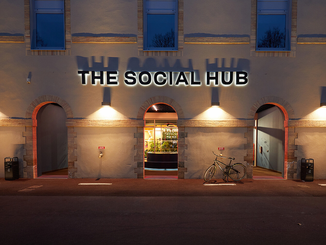 Facciata dell’edificio con tre ingressi ad arco illuminati al tramonto, con una bicicletta parcheggiata vicino a uno degli ingressi e un cartello con la scritta "THE SOCIAL HUB" sopra l’arco centrale