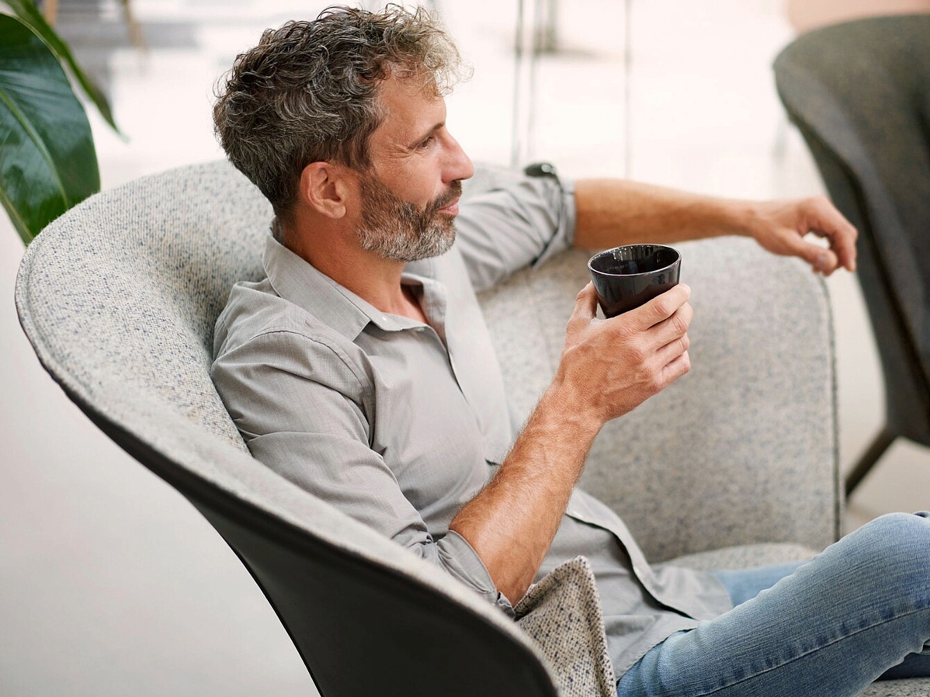 Mann mit grauem Hemd sitzt entspannt in einem hellgrauen Sessel und hält eine schwarze Tasse in der Hand