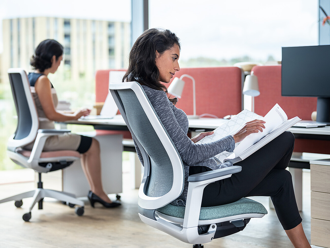 Two women working in a modern office, one reading documents and the other typing on a laptop