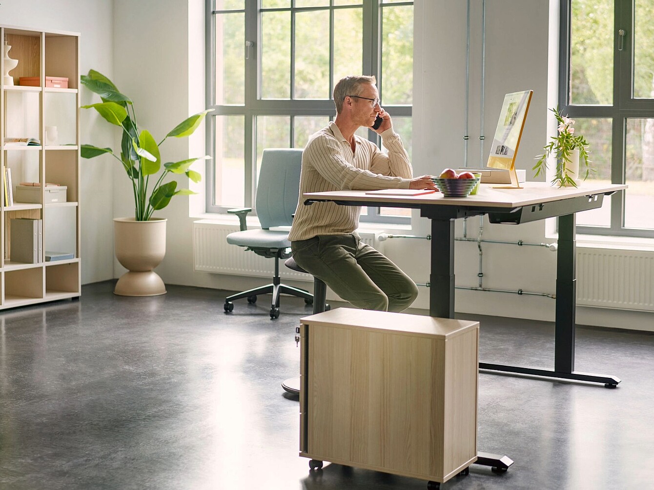 Persona sentada en una silla se:fit frente a un escritorio de altura regulable en una oficina luminosa con grandes ventanas y plantas