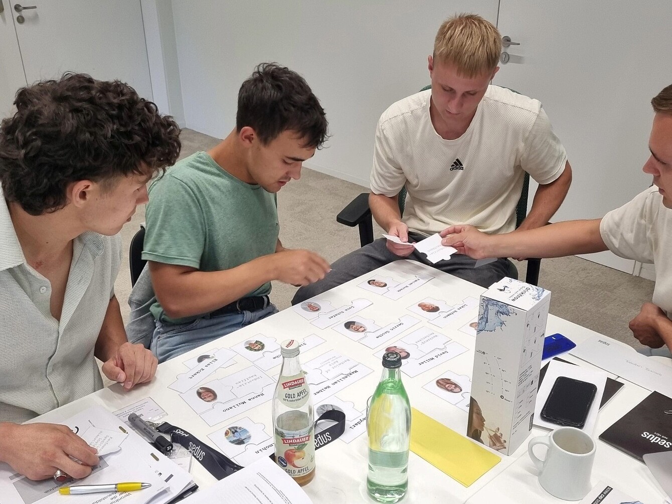 Cuatro jóvenes sentados alrededor de una mesa con tarjetas de visita, botellas, una taza de café, un smartphone y papeles, participando en una actividad en grupo en una sala sencilla