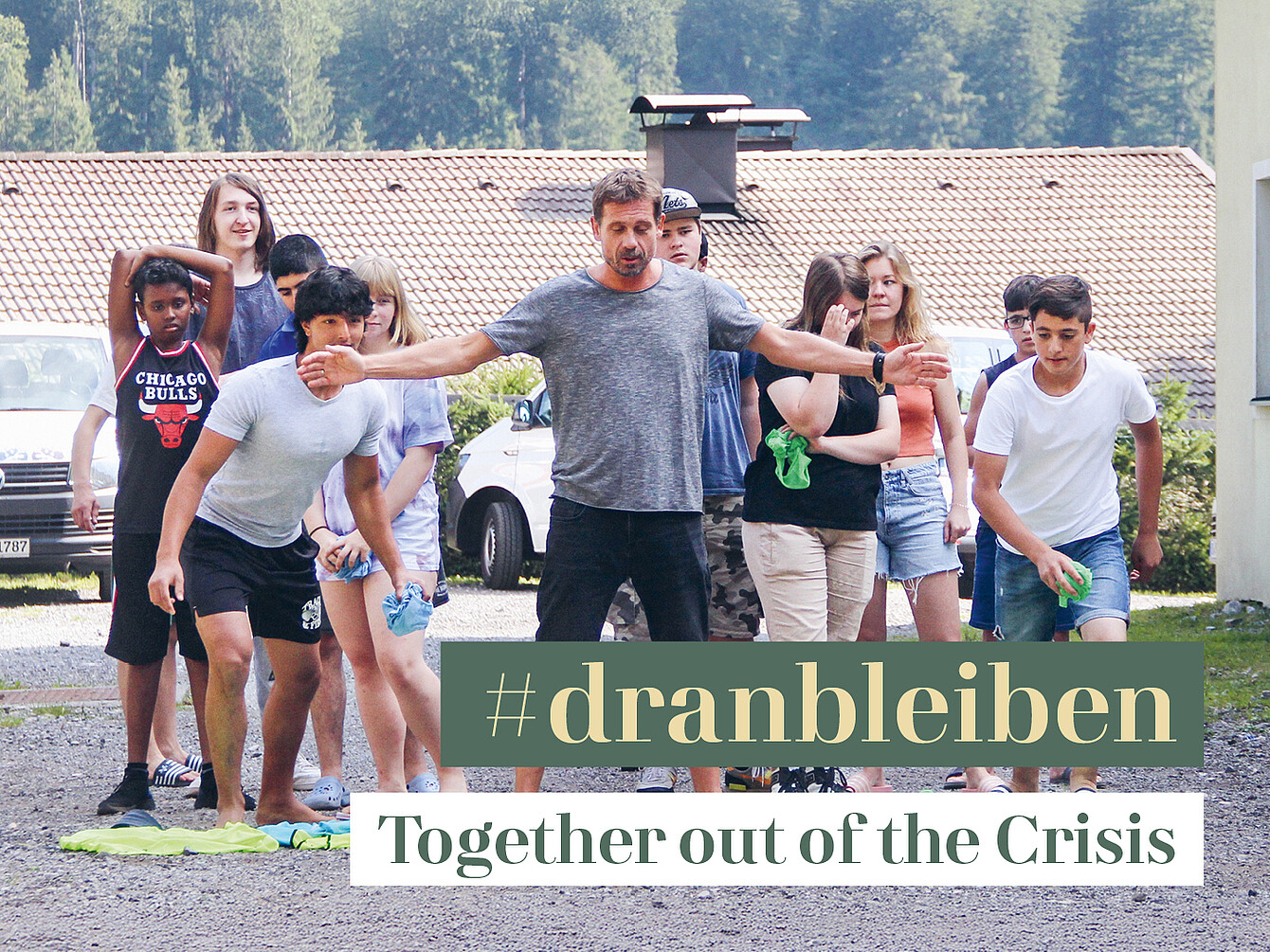 Grupo de jóvenes al aire libre, algunos haciendo estiramientos y otros de pie, con un edificio y coches aparcados al fondo, superpuesto con el texto «#dranbleiben Juntos para salir de la crisis».