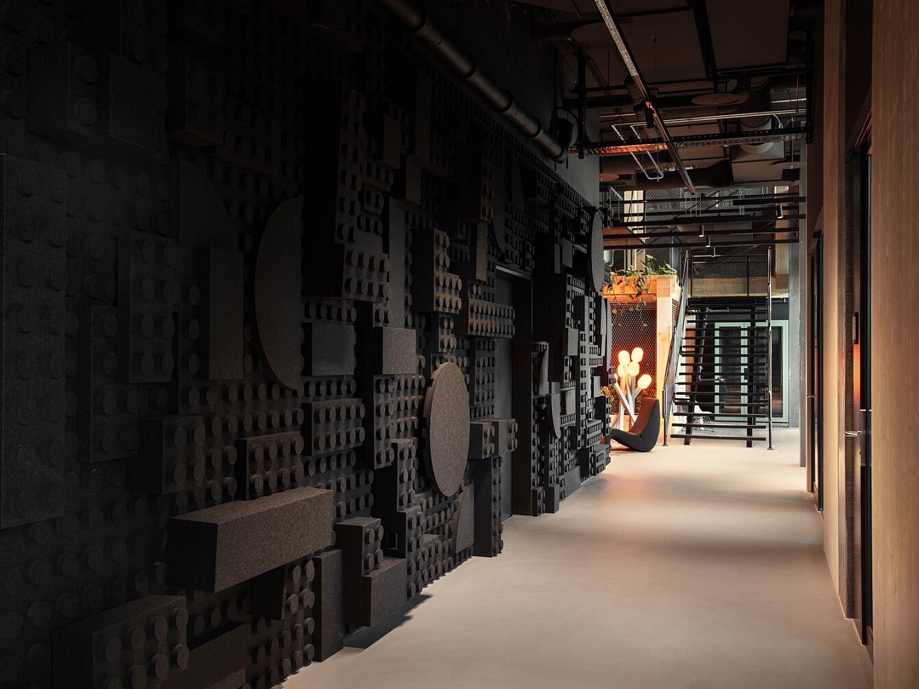 Long corridor with a dark wall featuring large geometric foam panels and a light floor, leading to a seating area with a person and a staircase.