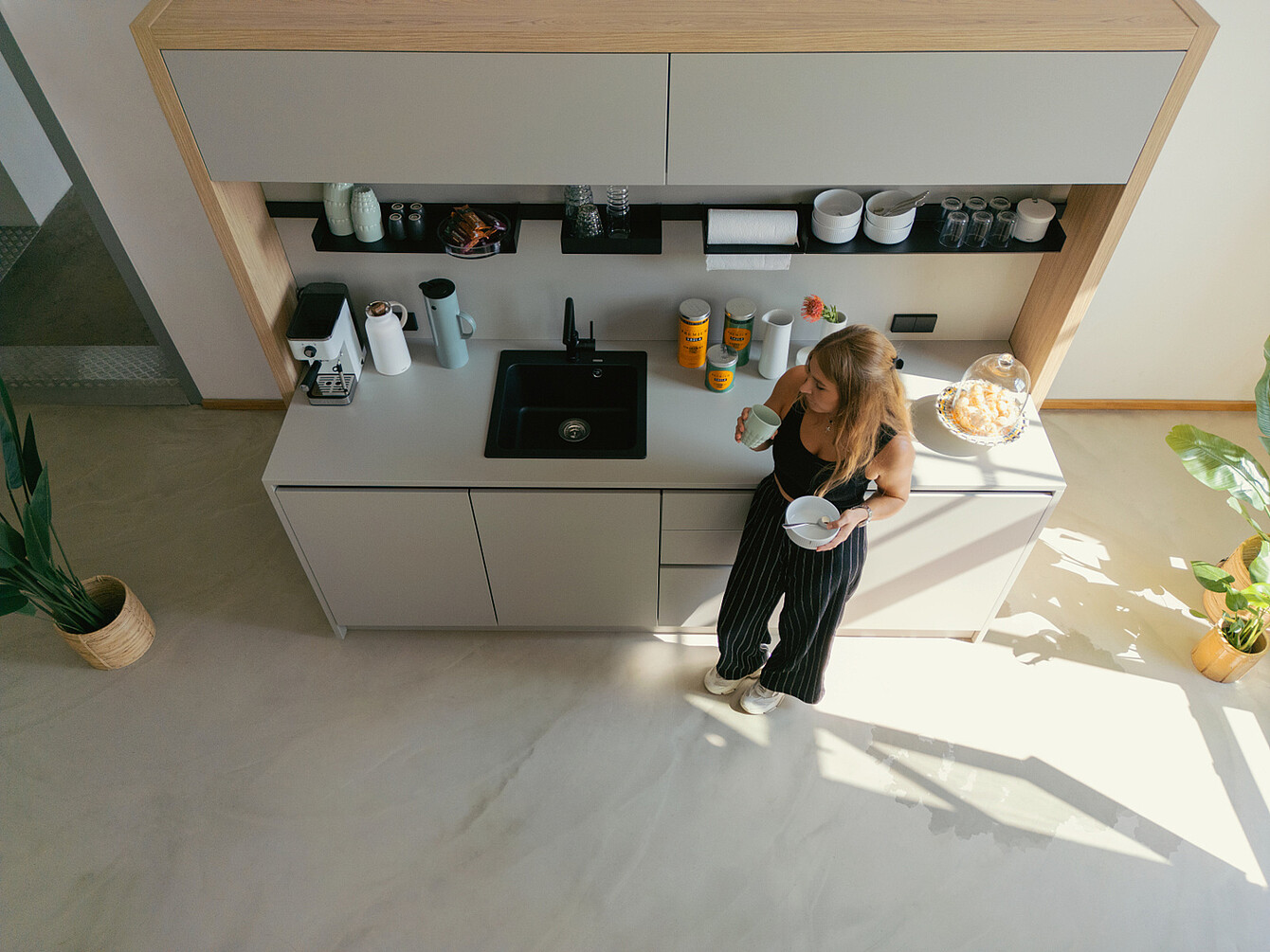 Donna in top nero e pantaloni a righe in piedi in una luminosa cucina dell'ufficio con luce naturale, con in mano una tazza e una ciotola vicino a un bancone con macchina del caffè, barattoli e piante.