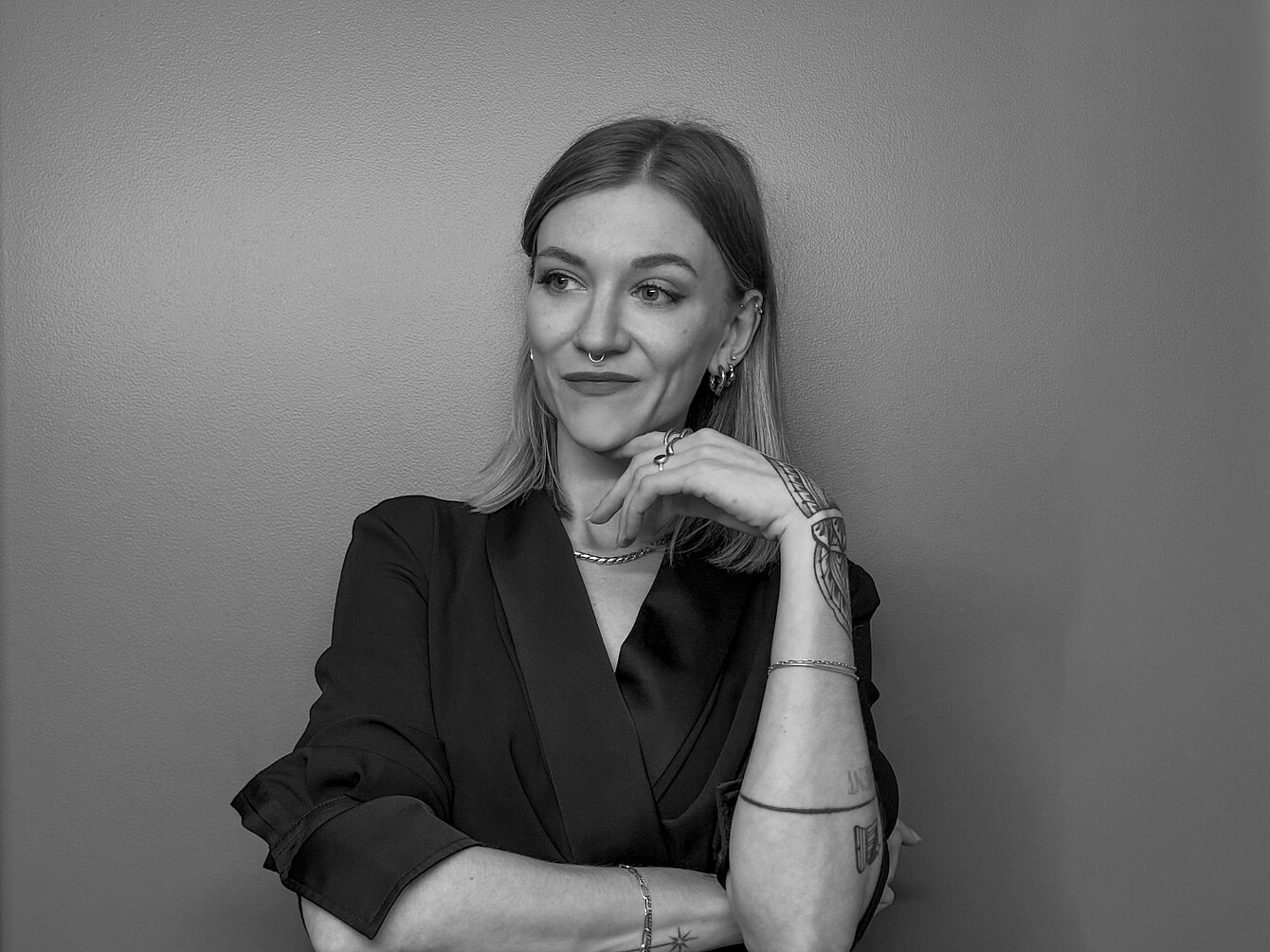 Person with shoulder-length hair wearing a dark blazer, resting chin on hand, showing multiple bracelets and tattoos on forearm.