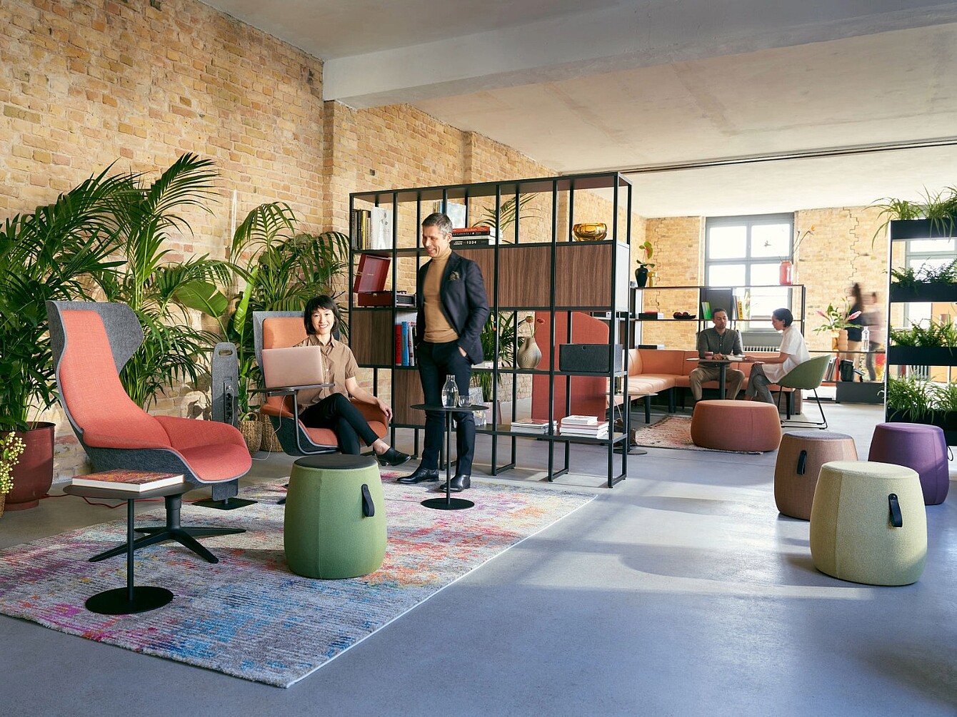Open lounge area with modern seating including armchairs, stools, and a modular sofa separated by a black metal and wood shelving unit, set against a brick wall with large windows and indoor plants