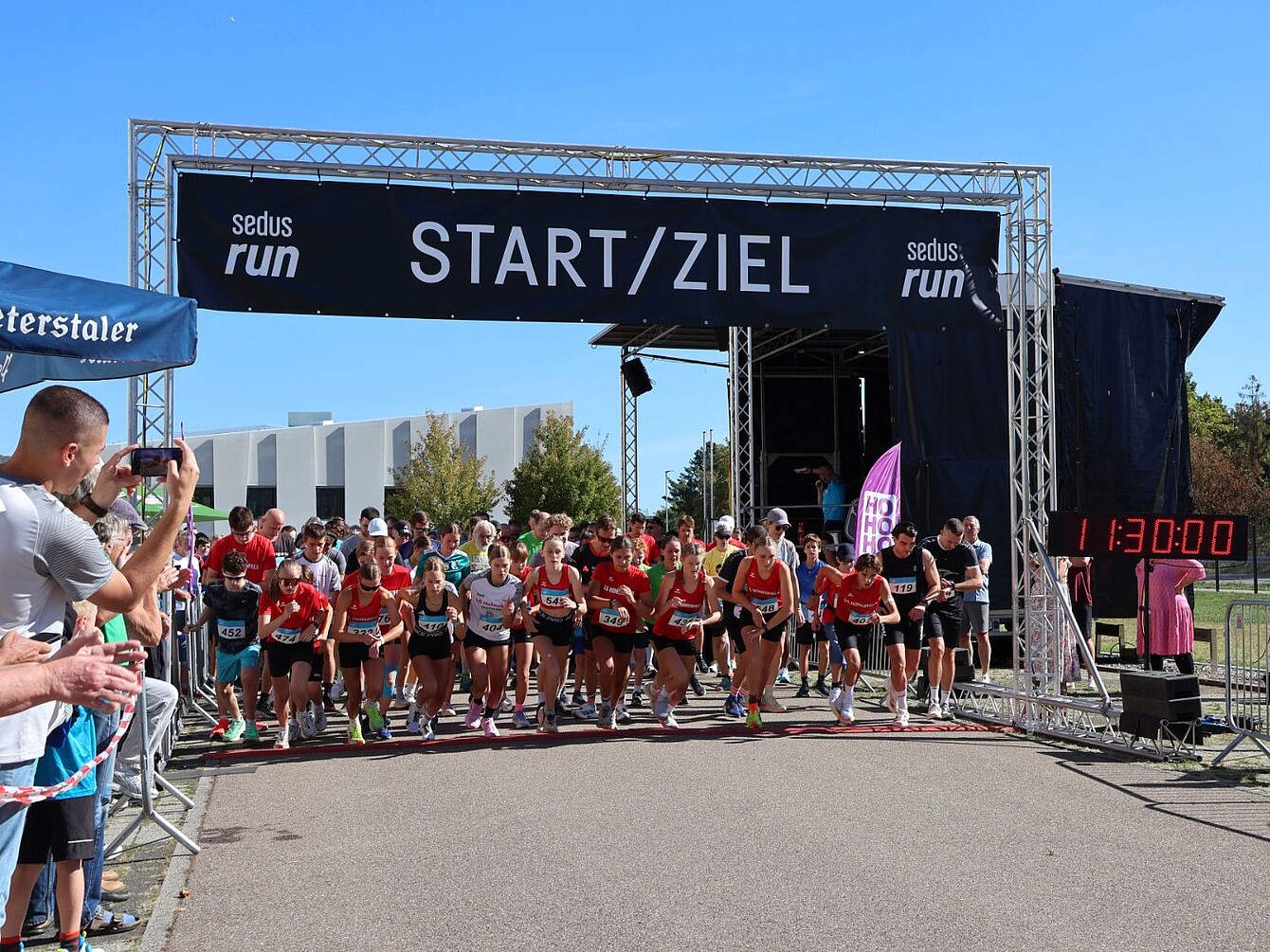 Startlinie eines Laufs mit vielen Läufern in Sportkleidung unter einem Banner mit der Aufschrift 'START/ZIEL' und digitaler Uhr mit der Zeit '11:30:00'