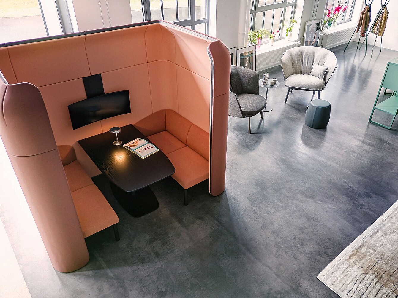 Cabina curvada con paredes altas, asientos tipo banco integrados y una mesa negra dentro de una sala abierta con dos sillones y una pequeña mesa redonda cerca.