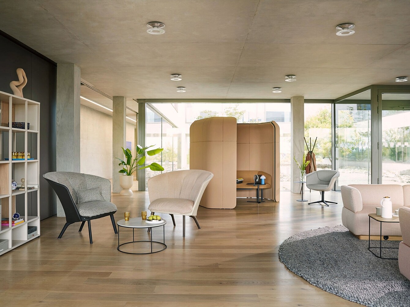 Moderner, offener Raum mit mehreren Sesseln unterschiedlicher Formen und Farben, einem kleinen runden Tisch, einem grauen Teppich und einer halbhohen, beige gepolsterten Trennwand im Hintergrund