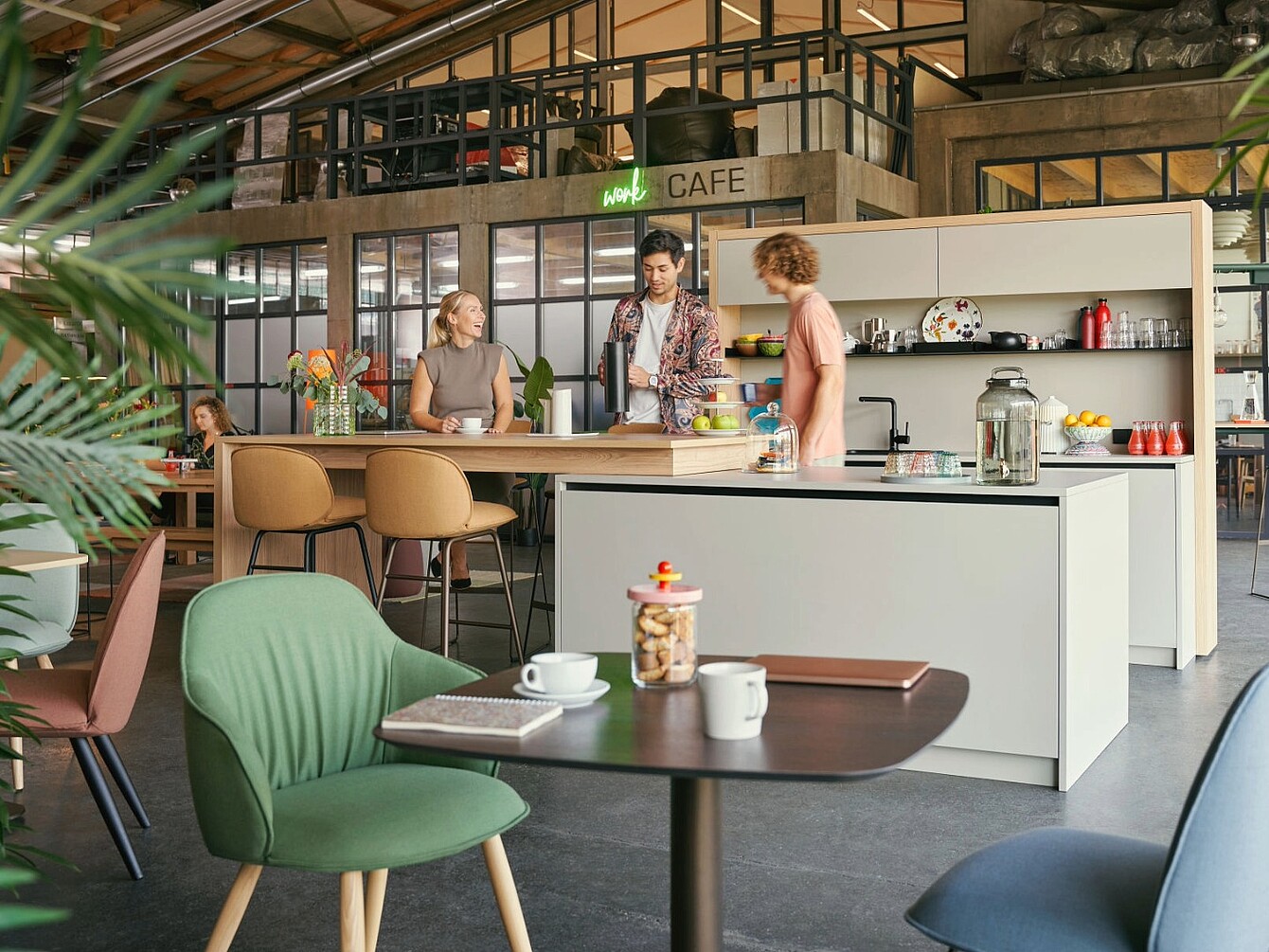 Three people standing and talking around a kitchen island in a modern office cafe area with seating and plants