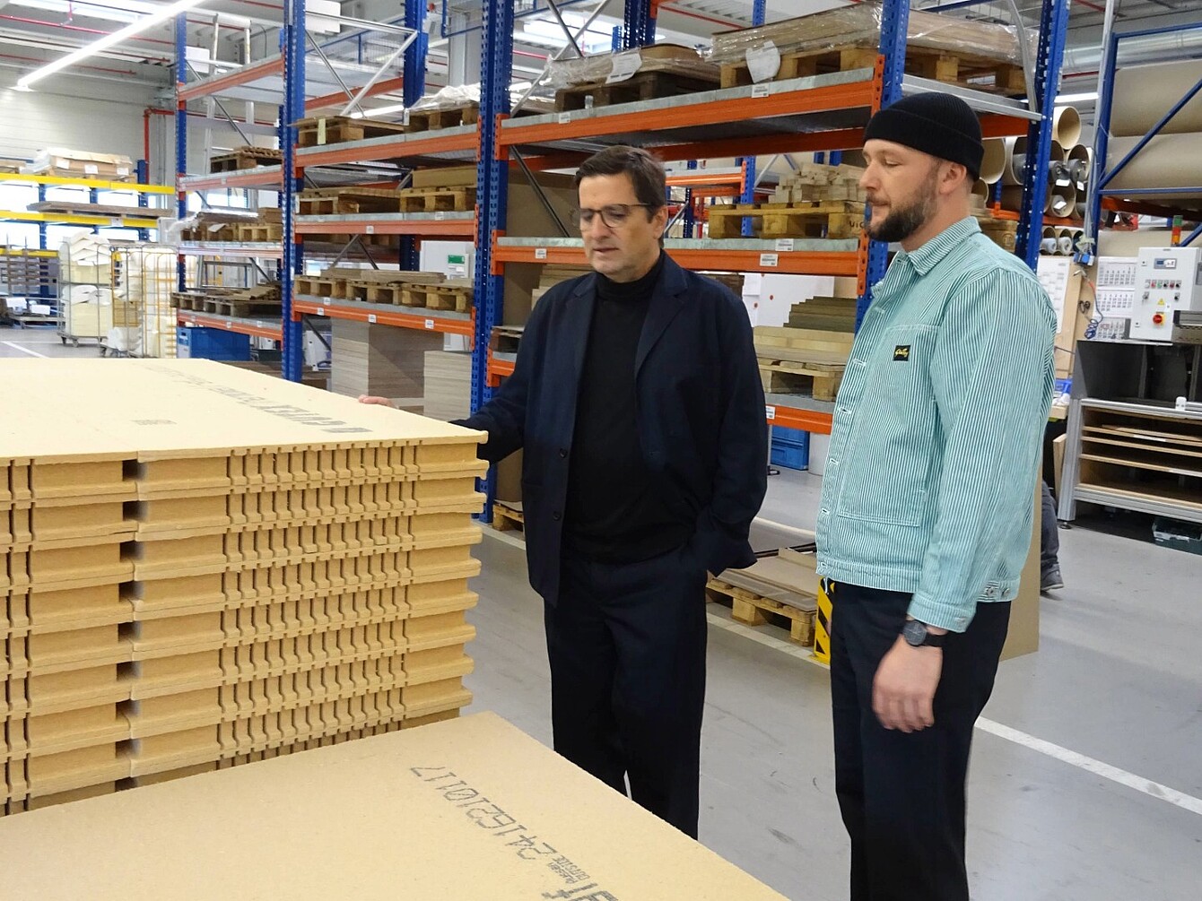 Two men (Rudi Raschke and Konstantin Thomas) stand beside stacks of wood fibre panels in a warehouse with metal shelving and industrial equipment.