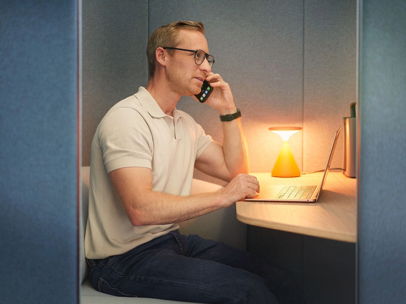 Mann mit Brille sitzt in einem kleinen Büro mit blauen Wänden, telefoniert und arbeitet an einem Laptop, auf dem Tisch steht eine orangefarbene Lampe
