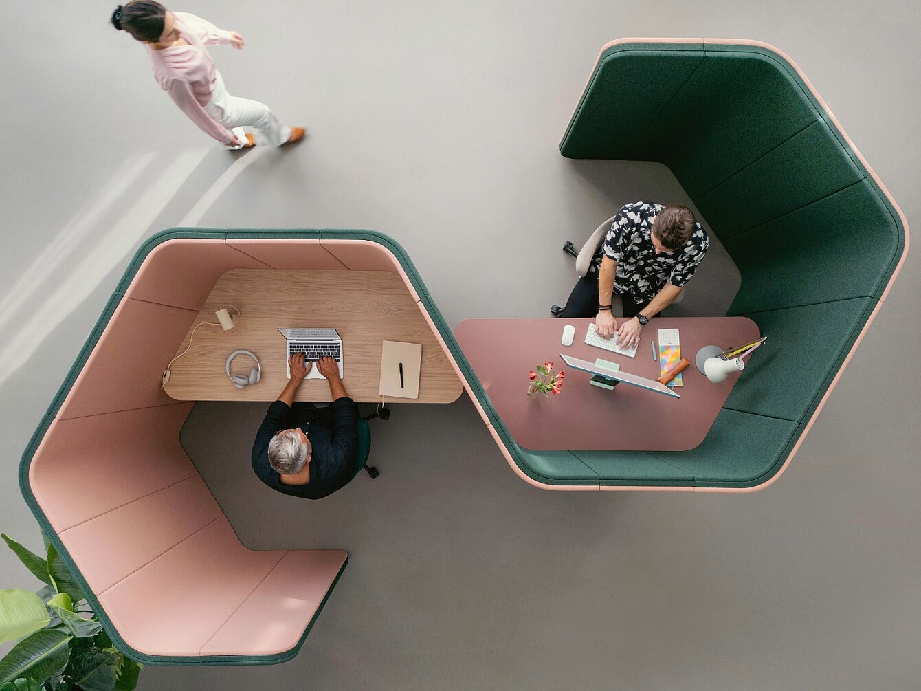 Two people working separately at curved desks with high upholstered partitions in an open office space, one typing on a laptop and the other writing on paper, while a third person walks nearby.