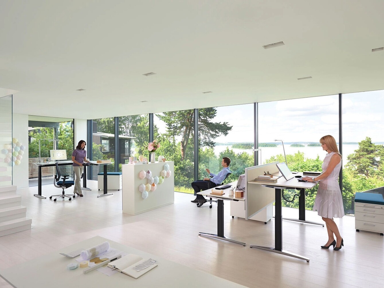 Modern office space with large windows showing a green outdoor view, featuring three people using height-adjustable desks in sitting and standing postures, with minimalistic white furniture and light flooring.