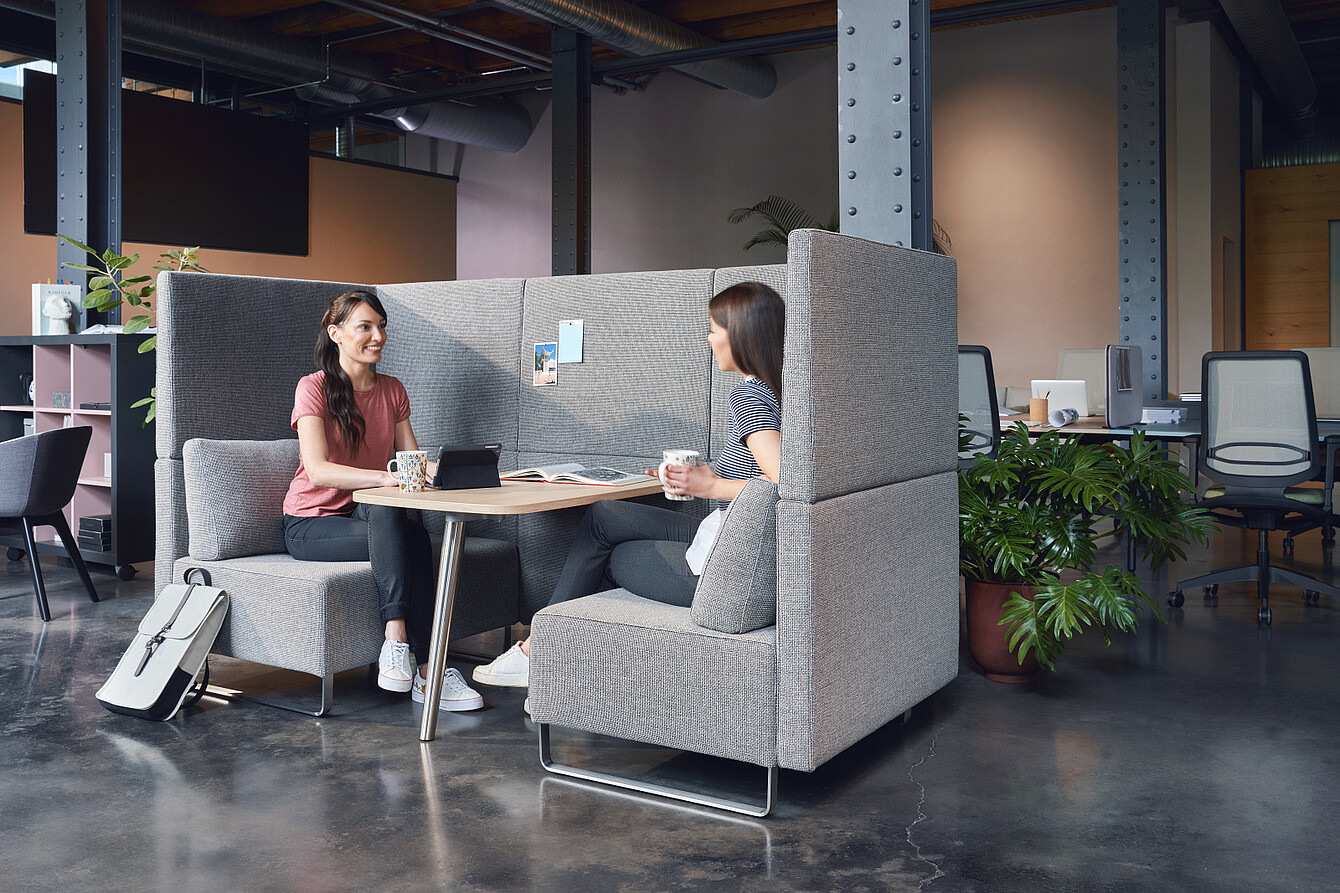 Zwei Frauen sitzen sich in einer grauen, hochwandigen Lounge-Ecke gegenüber und tauschen sich aus