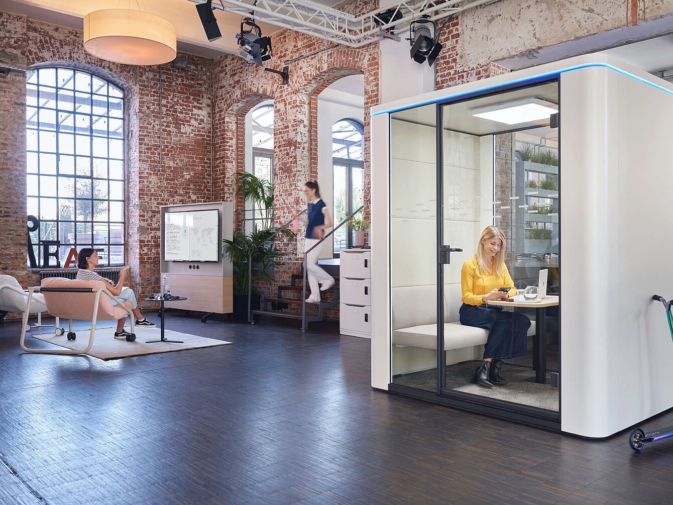 Modern office space with exposed brick walls and large windows featuring a woman sitting inside a glass-enclosed acoustic pod working on a laptop and two other women in the open area, one seated on a chair and the other walking up stairs