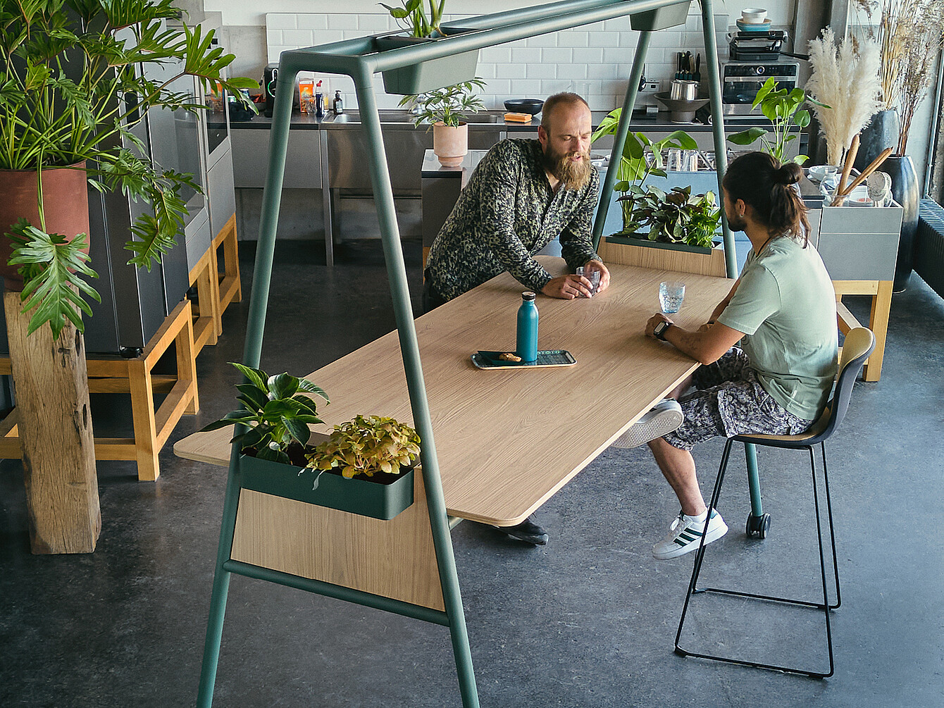 Two men having a discussion at a wooden table with integrated planters in a modern workspace designed to inspire focused work and collaborative creativity, surrounded by plants and kitchen elements.