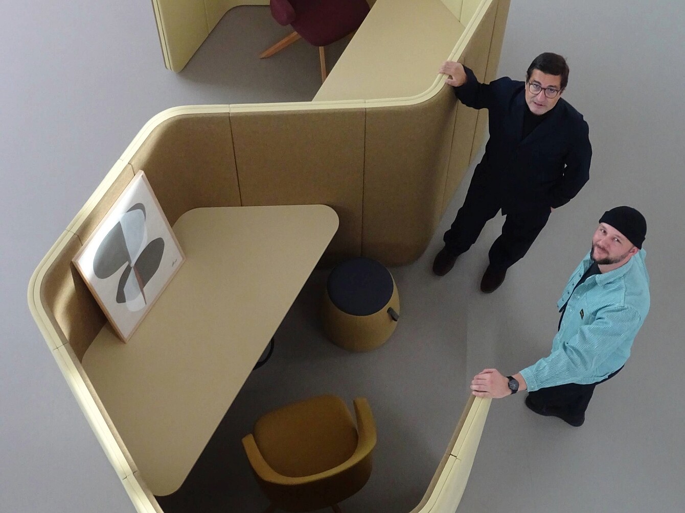 Two men (Rudi Raschke and Konstantin Thomas) stand beside a curved, beige partitioned workspace with a mounted abstract artwork, a mustard-yellow chair, and a small round stool inside.