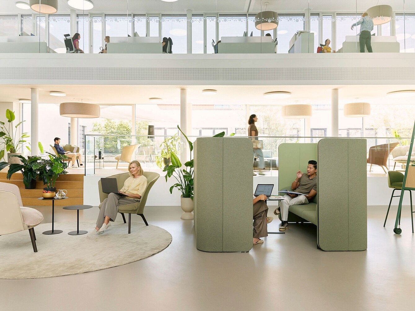 Interior de oficina moderno con zonas de asientos abiertas, cabinas de privacidad tapizadas en verde, plantas y grandes ventanas que proporcionan luz natural