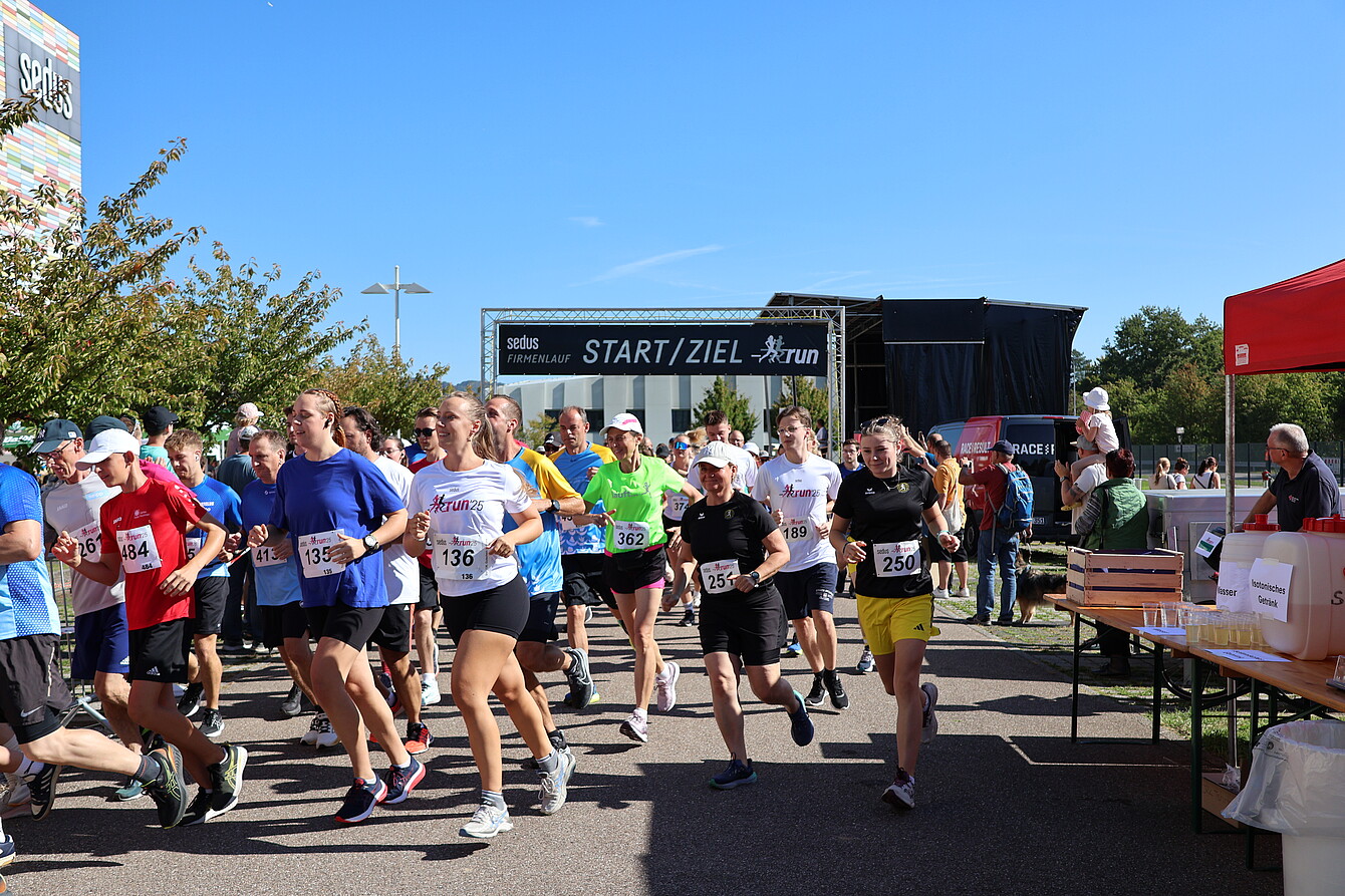 Runners starting the Sedus RUN under a black banner reading 'START/ZIEL run' on a sunny day, with participants wearing numbered bibs and athletic clothing.