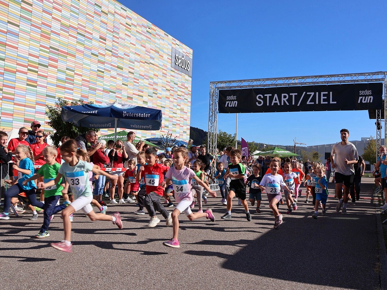 Kinder starten einen Lauf vor einem Start-Ziel-Banner mit der Aufschrift 'sedus run START/ZIEL' vor einem bunten Gebäude und Zuschauern im Hintergrund