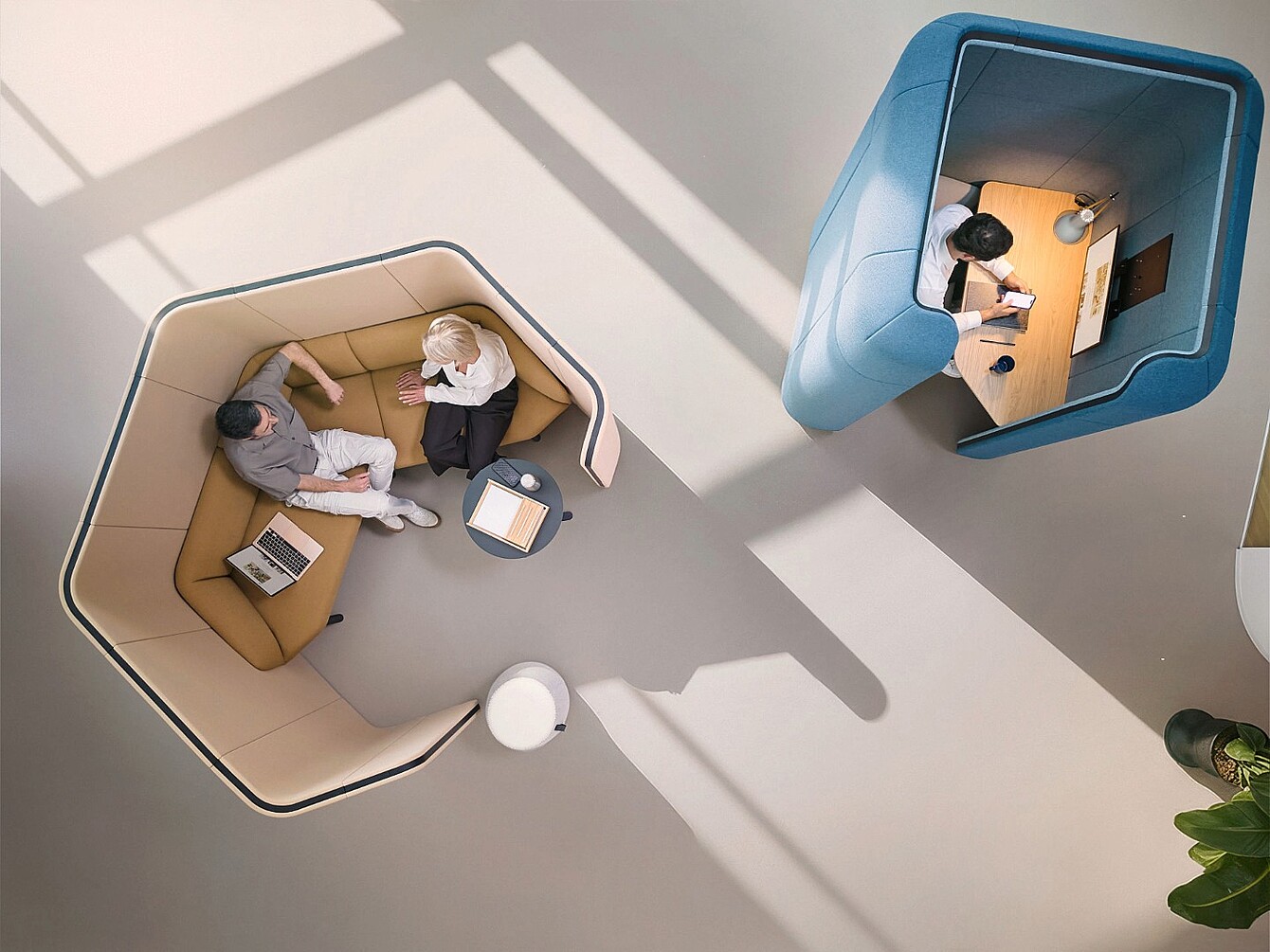 Two people sitting in a beige soundproof office pod with a small table and laptop opposite a person working alone in a blue soundproof office pod with a desk and laptop