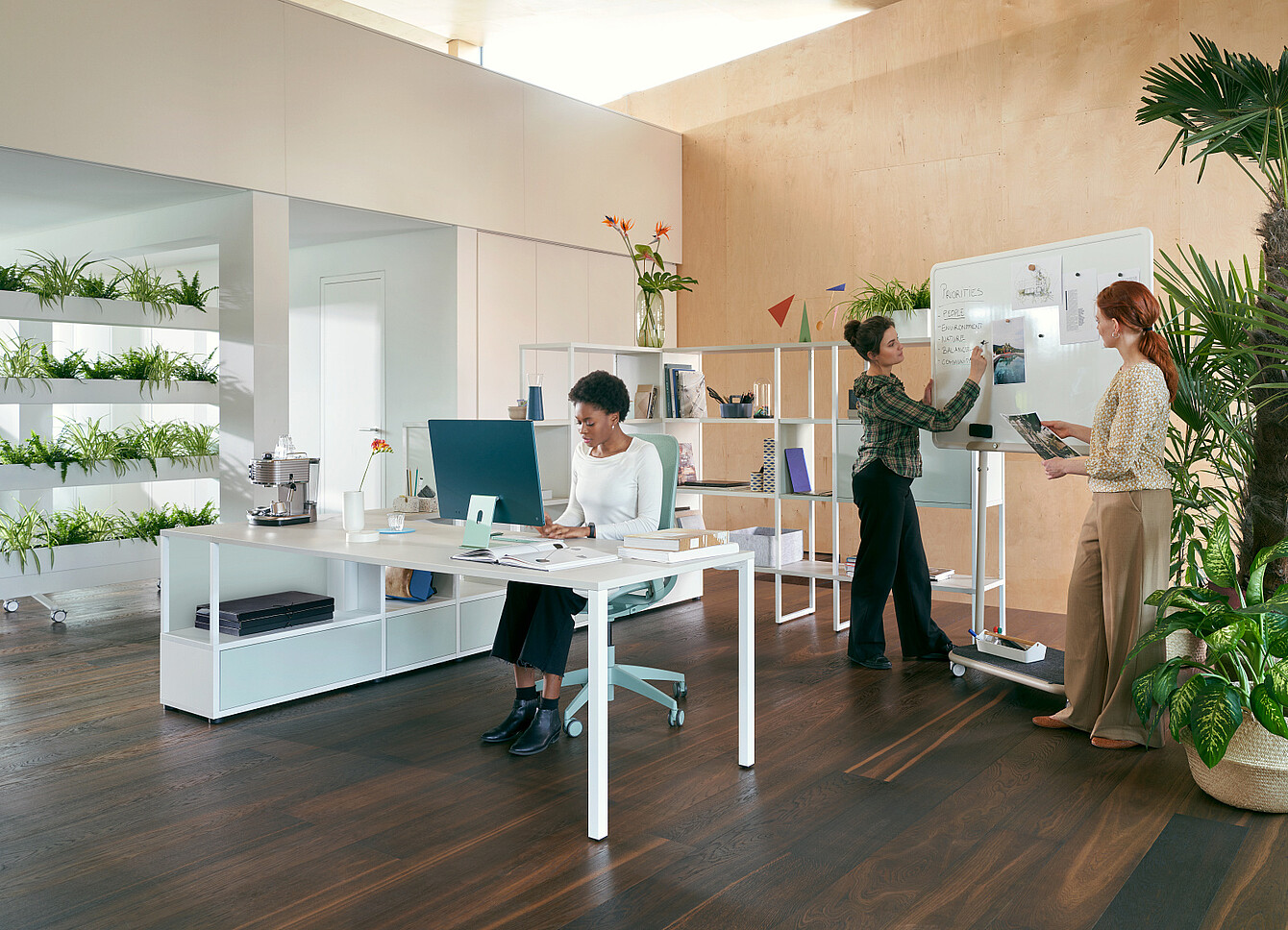 Drei Frauen in modernem Büro, eine sitzt am Schreibtisch vor Computer, zwei stehen vor Whiteboard mit Notizen und Tablet