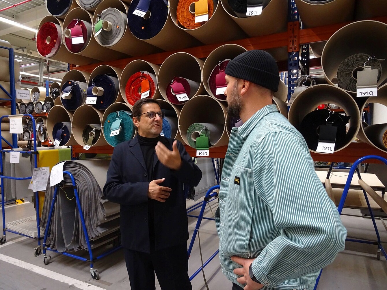 Two men (Rudi Raschke and Konstantin Thomas) stand and talk in a warehouse aisle with large rolls of fabric or material stored on shelves behind them.