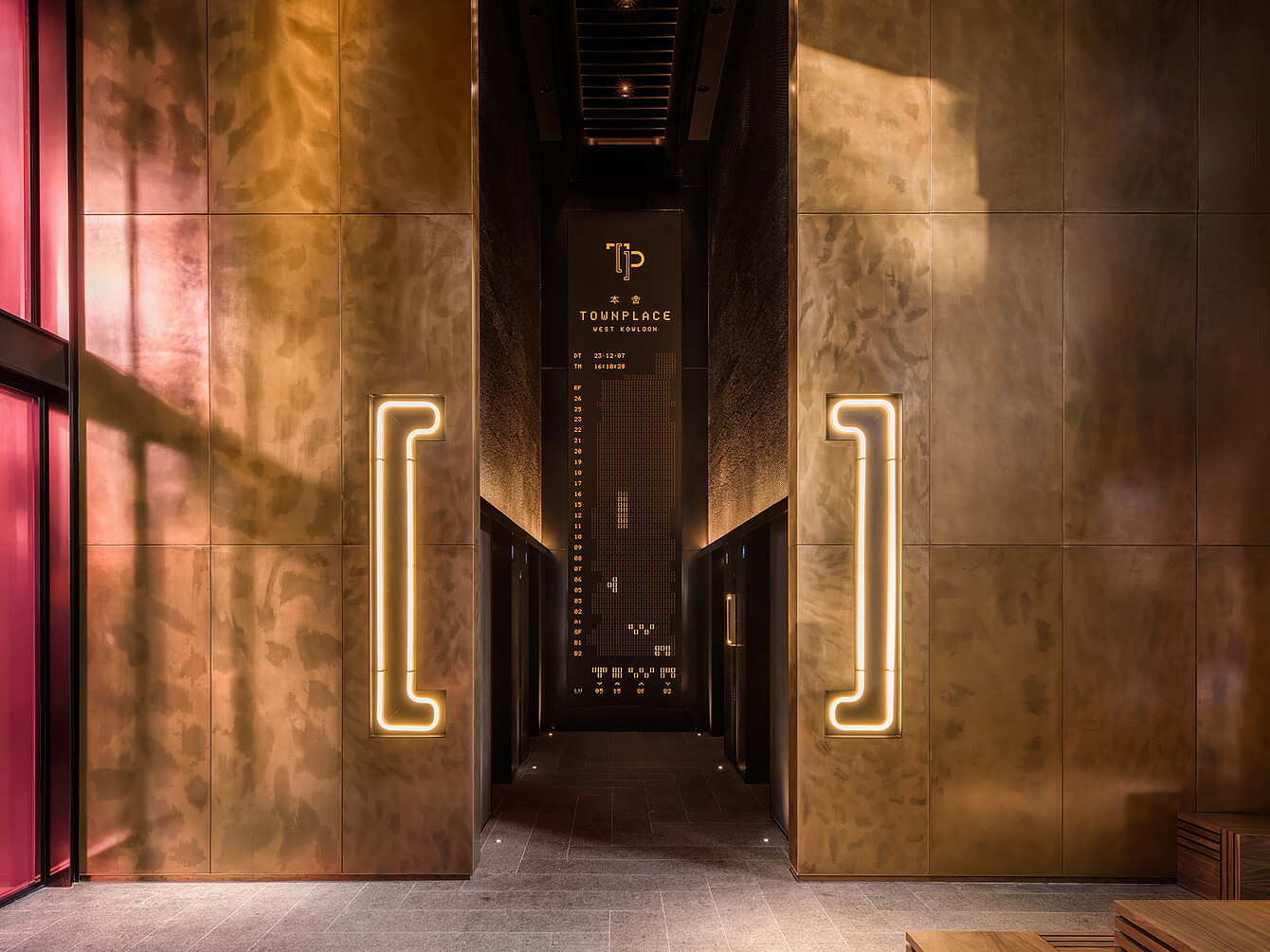 Stretto corridoio con pareti color bronzo e neon a forma di parentesi che conduce a un ingresso buio con un led con la scritta "TP TOWNPLACE"