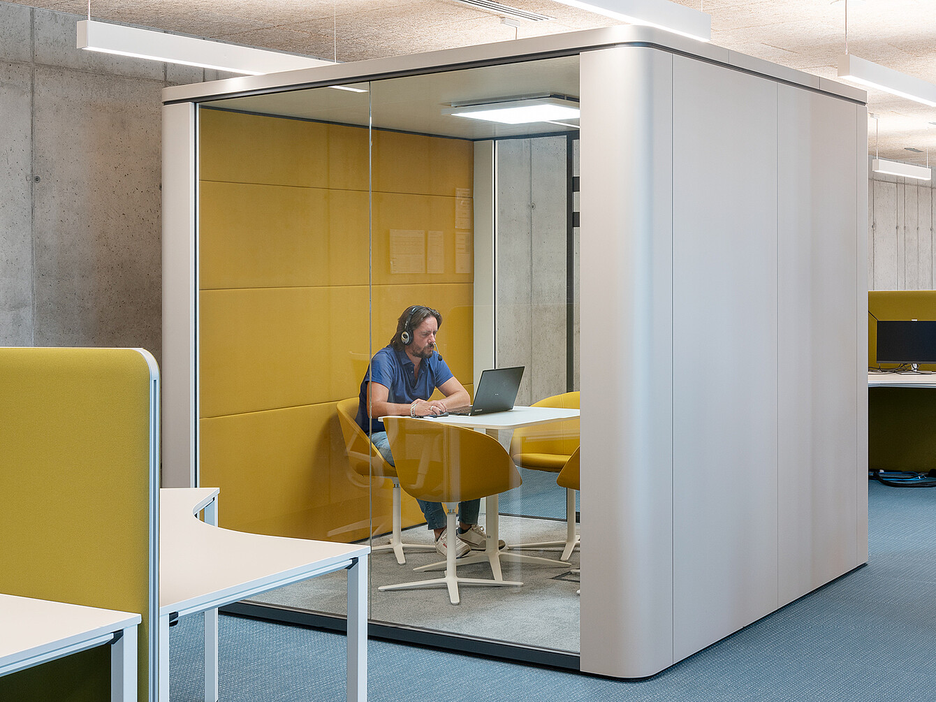 Person wearing headphones sitting at a table with a laptop inside a glass-walled office pod with yellow padded walls and chairs