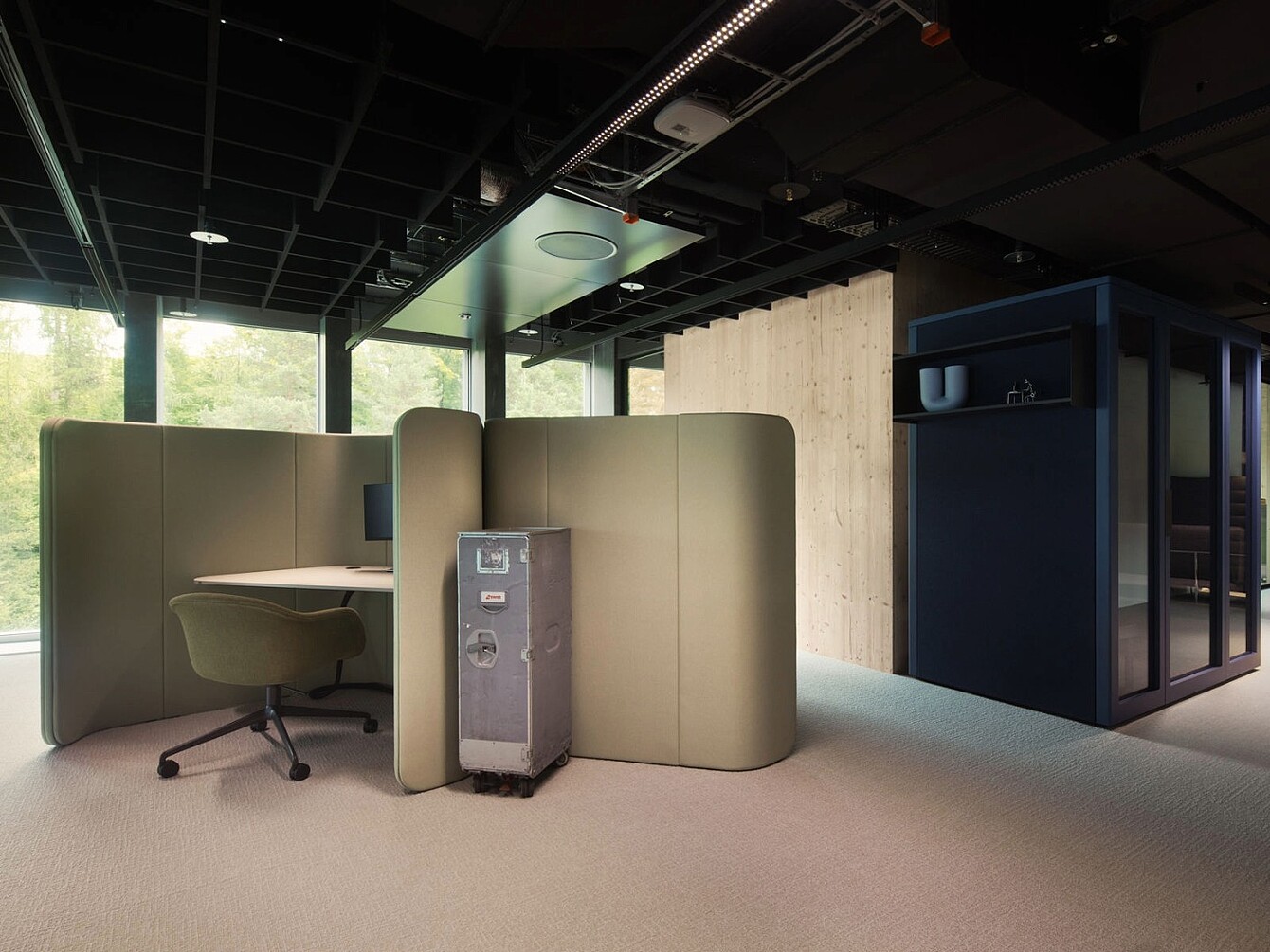 Work area with a beige curved partition enclosing a desk and an olive-green swivel chair, next to a metal storage box and a dark blue shelving unit with cups.