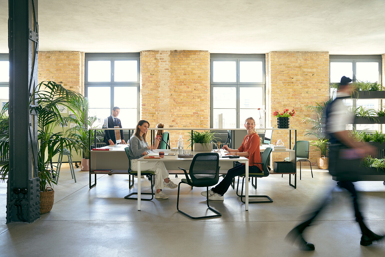 Offenes Büro mit großen Fenstern, zwei sitzenden Frauen an Tischen und einer vorbeigehenden Person im Vordergrund