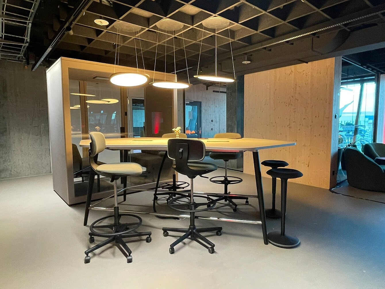 High table with multiple adjustable swivel stools around it in a modern office space at Novu Campus, Zurich Airport, with pendant lights overhead and wooden walls.