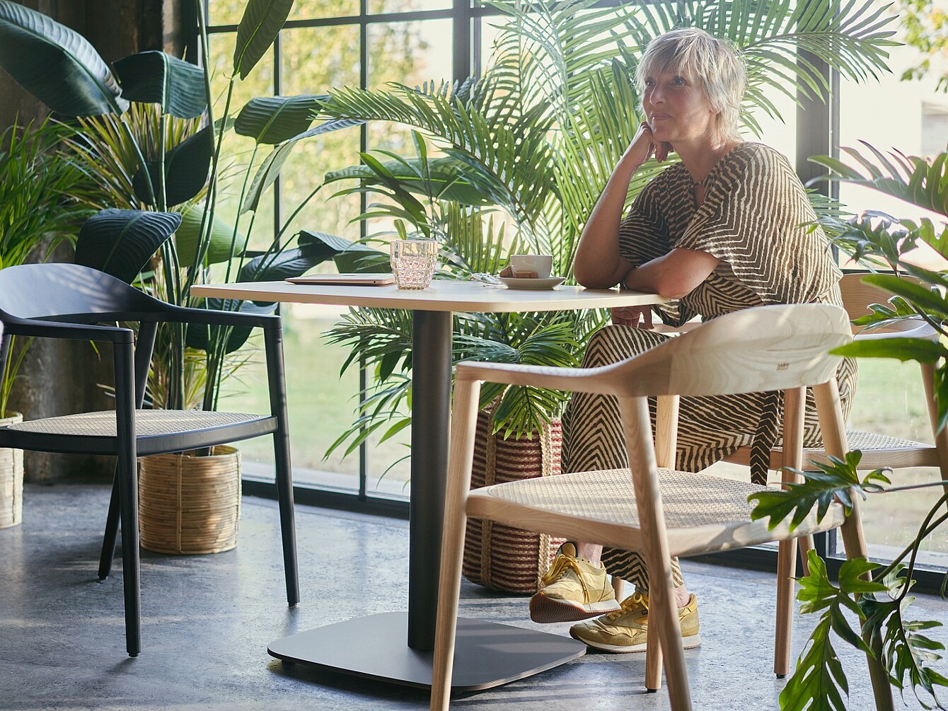 Person sitting at a table in a room with large windows and multiple office plants around