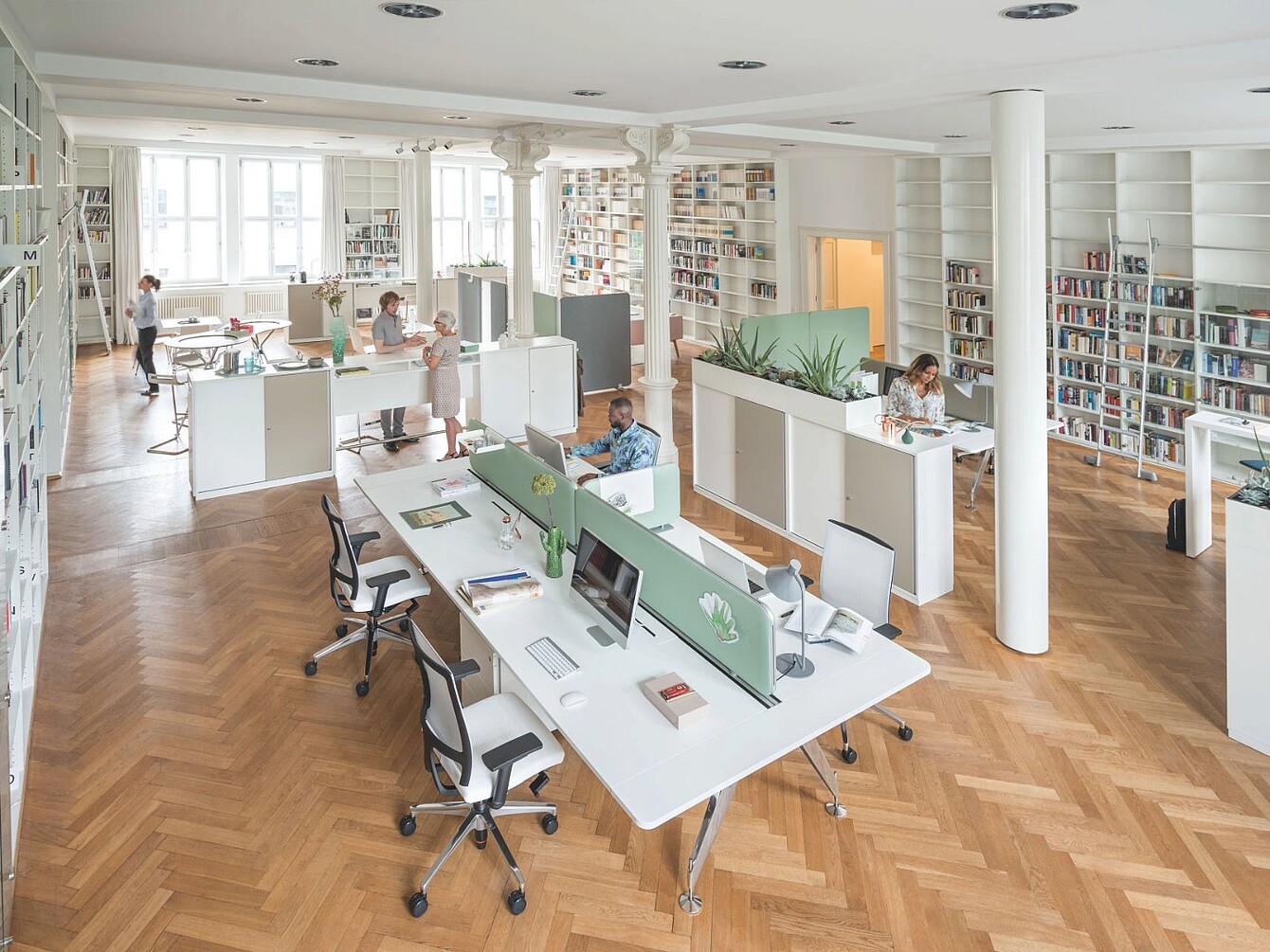 Open-plan office with desks separated by green fabric panels, multiple people working, bookshelves lining the walls, and wooden parquet flooring