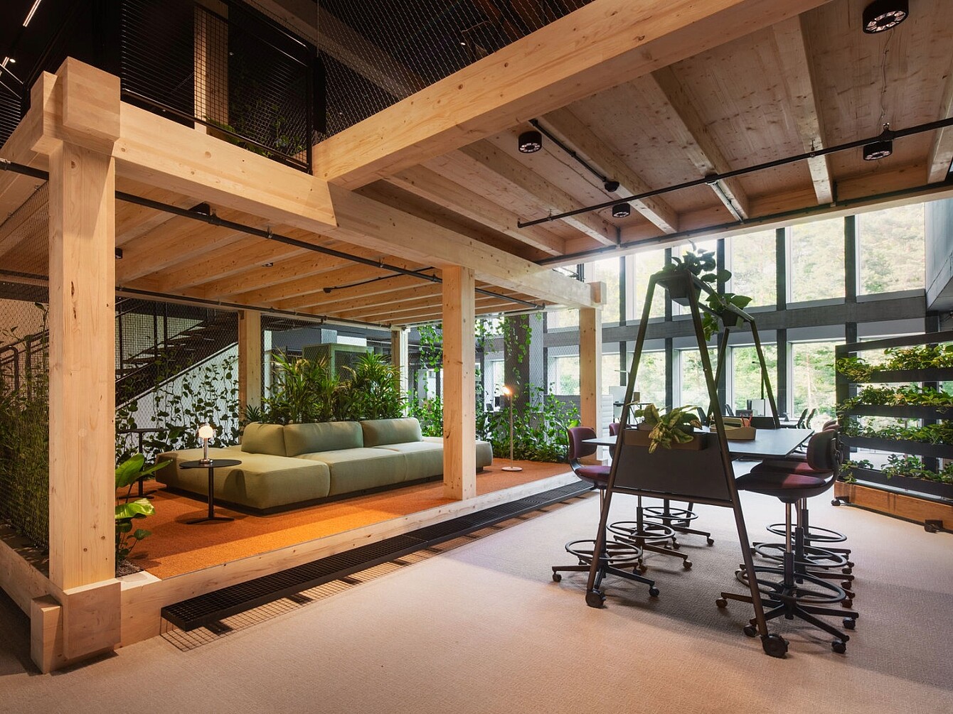 Open office area with wooden beams, green sofa, multiple chairs around a table, and plants near large windows.