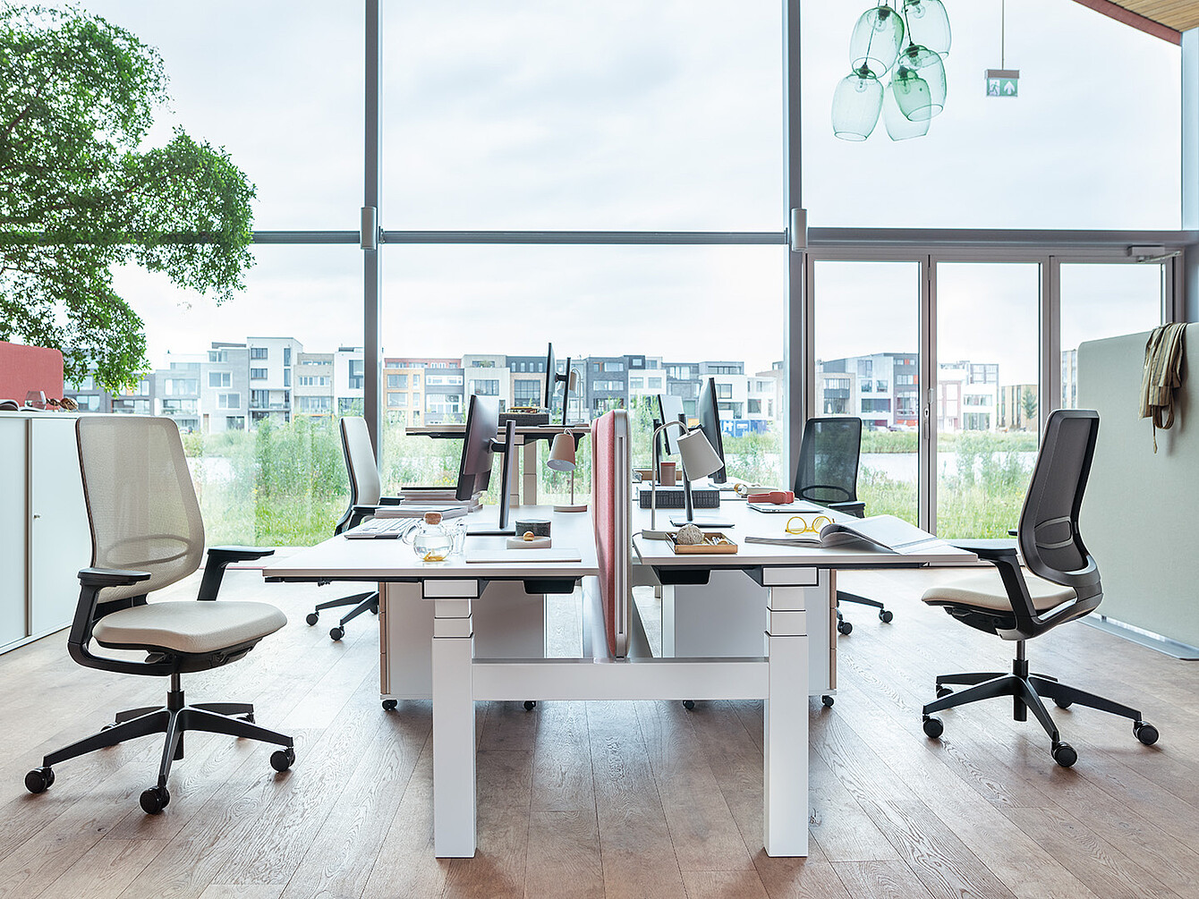 Modern office workspace with four desks arranged in two rows facing each other, ergonomic chairs, computer monitors, and large windows showing an urban view outside