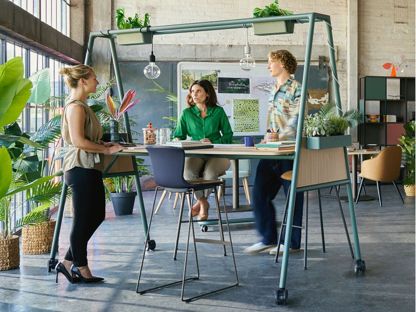 Three people around a high table with plants on and around it in a spacious office with large windows and exposed brick walls